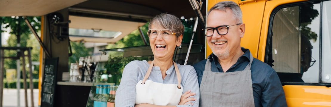 Retired couple in aprons smiling beside their food truck, starting a small business together in retirement.